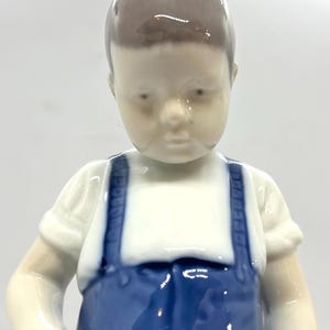 May include: A glazed porcelain figurine of a child with short brown hair, wearing a white short-sleeved shirt and blue overalls with shoulder straps. The figure has a smooth, glossy finish and is depicted from the chest up against a plain background.
