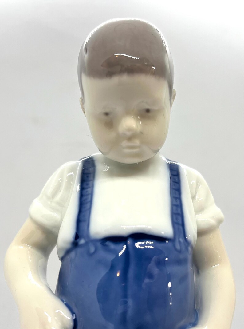 May include: A glazed porcelain figurine of a child with short brown hair, wearing a white short-sleeved shirt and blue overalls with shoulder straps. The figure has a smooth, glossy finish and is depicted from the chest up against a plain background.