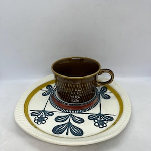 Puede incluir: Una taza de té de cerámica marrón con asa descansa sobre un platillo a juego. El platillo tiene un fondo blanco con un diseño floral azul y un borde dorado. El juego de taza y platillo es de estilo vintage.