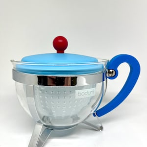 May include: A blue glass teapot with a stainless steel strainer and a red knob on the lid. The teapot has a blue silicone handle and sits on three metal legs. The word "bodum" is printed on the side of the teapot.