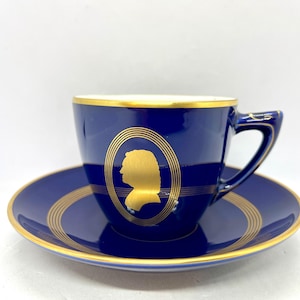 May include: A blue and gold teacup and saucer set with a gold silhouette of a person's profile on the cup.