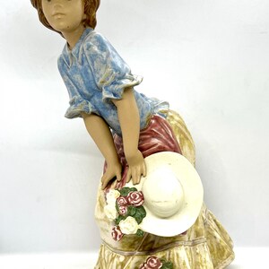 May include: A porcelain figurine of a girl wearing a blue shirt, a red and white skirt, and a white hat. She is holding a bouquet of roses.