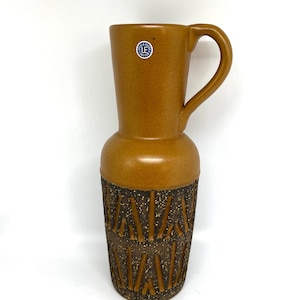 May include: A tall, brown ceramic pitcher with a handle. The upper portion is smooth, while the lower section features a textured, geometric design. A small, circular sticker is affixed near the handle. The pitcher is likely vintage or retro.