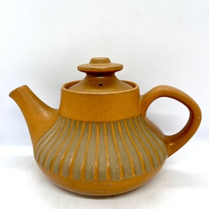 Może przedstawiać: Brązowy ceramiczny czajnik z żebrowanym wzorem i pokrywką. Czajnik ma dzióbek i uchwyt.