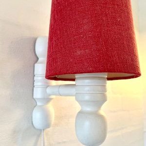 Może przedstawiać: Biała lampa ścienna z czerwonym abażurem z tkaniny. Lampa ma prosty design z dwoma białymi drewnianymi ramionami i okrągłą podstawą.