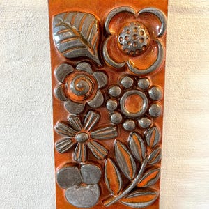 May include: An orange ceramic tile with a raised floral design. The flowers and leaves are outlined in silver and have orange accents. The tile features various flower shapes and leaf patterns. The tile measures approximately 15 cm x 30 cm.
