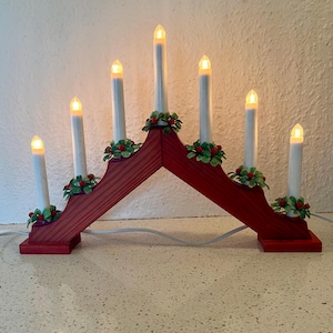 Puede incluir: Un puente de velas arqueado de madera roja con siete velas cónicas eléctricas blancas. Cada vela está iluminada con un brillo cálido y decorada en la base con hojas de acebo verdes y pequeñas bayas rojas. Incluye un cable de alimentación blanco.