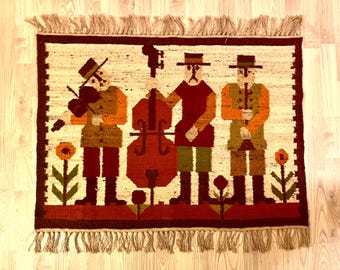 May include: A woven wall hanging featuring three figures in brown, red, and green clothing playing musical instruments. The figures are surrounded by stylized flowers and plants. The background is a cream color.