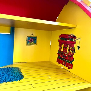 Op de afbeelding: Kleurrijk houten poppenhuisinterieur met een rood dak, gele muren en een blauwe deur. Het interieur heeft een klein vloerkleed, een schilderij van een schip en decoratieve stickers. De vloer is geschilderd om op houten planken te lijken.