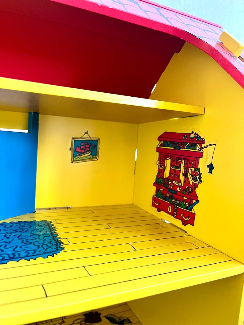 Op de afbeelding: Kleurrijk houten poppenhuisinterieur met een rood dak, gele muren en een blauwe deur. Het interieur heeft een klein vloerkleed, een schilderij van een schip en decoratieve stickers. De vloer is geschilderd om op houten planken te lijken.