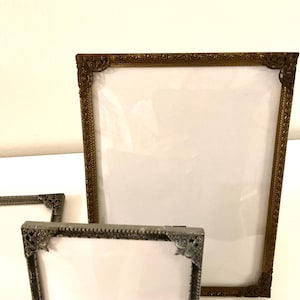 May include: Three ornate picture frames in varying sizes. The largest frame is bronze-colored, while the smaller frames are silver-toned. All frames have decorative corner details and a white backing, ready for photos or artwork.