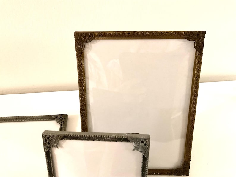 May include: Three ornate picture frames in varying sizes. The largest frame is bronze-colored, while the smaller frames are silver-toned. All frames have decorative corner details and a white backing, ready for photos or artwork.