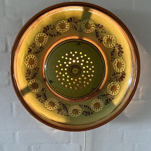 May include: A circular, green ceramic wall light with a brown border. The light has a floral pattern around the edge and a perforated centre. It is mounted on a white brick wall and has a white power cable.