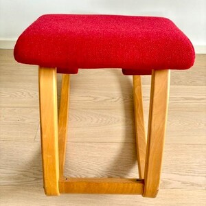 May include: A small stool with a red upholstered seat. The stool has a rectangular seat and four wooden legs. The legs are angled outwards. The frame and legs are made of light-colored wood. The seat is made of fabric.
