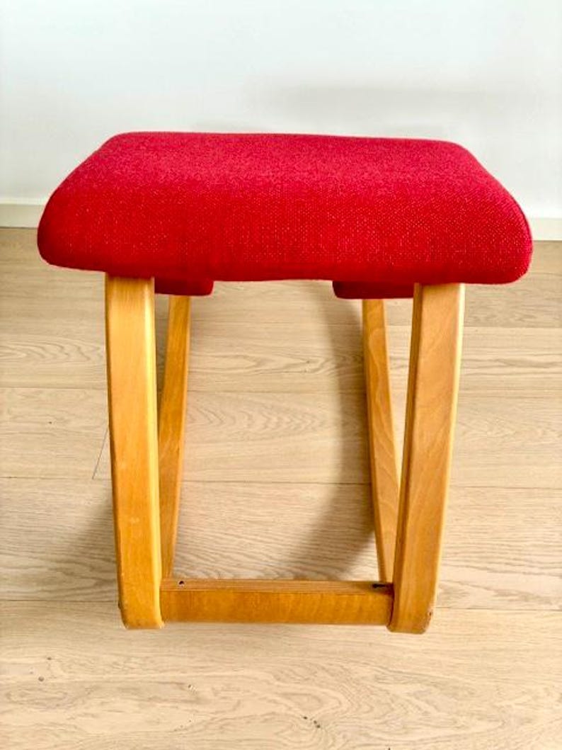 May include: A small stool with a red upholstered seat. The stool has a rectangular seat and four wooden legs. The legs are angled outwards. The frame and legs are made of light-colored wood. The seat is made of fabric.