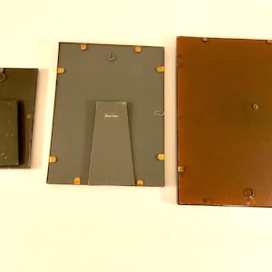 May include: Three rectangular picture frames of varying sizes, viewed from the back. The frames have dark gray and brown backing with brass-colored fasteners. Each frame has a built-in easel for tabletop display.