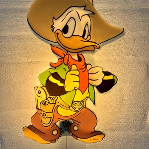 May include: A cartoon wall light shaped like a cowboy duck. The light has a tan cowboy hat, orange neckerchief, and brown boots. The character is holding a gun and wearing a green vest. The light is plugged into a wall.