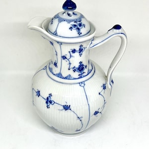 May include: A white porcelain pitcher with a lid, featuring a delicate blue floral pattern painted across the ribbed surface. The pitcher has a curved handle, a pointed spout, and a matching lid with a small blue finial on top.
