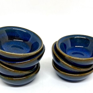 Peut inclure: Deux piles de petits bols en céramique ronds présentant un vernis intérieur bleu brillant profond et un bord ainsi qu'un extérieur brun terreux contrastant. Les bols ont une forme lisse et incurvée et sont disposés en deux piles distinctes.