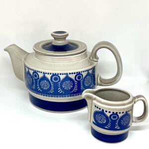 May include: A ceramic teapot and matching creamer featuring a light gray base with a vibrant blue band around the bottom and a repeating blue graphic pattern of stylized pomegranate-like shapes with circular details around the center.