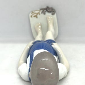 May include: A porcelain figurine of a person lying on their stomach, wearing a white shirt and blue shorts. The figure rests on a rectangular base decorated with a small brown crab and a tan coral-like design, viewed from above.