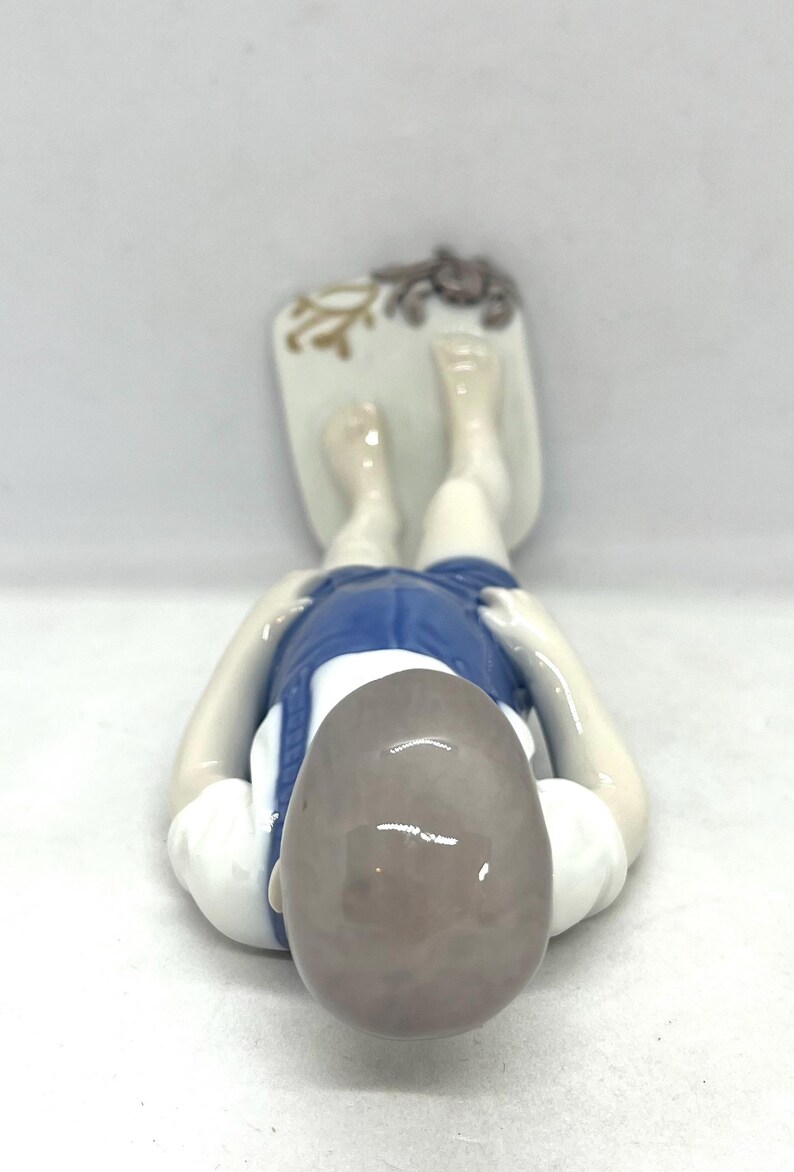 May include: A porcelain figurine of a person lying on their stomach, wearing a white shirt and blue shorts. The figure rests on a rectangular base decorated with a small brown crab and a tan coral-like design, viewed from above.