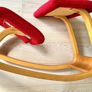 May include: A red kneeling chair with a curved wooden frame. The chair has two padded red cushions and a natural wood finish. The chair is designed to promote good posture and is suitable for use at a desk or table.