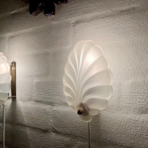 Könnte beinhalten: Zwei Wandleuchten mit Muscheldesign. Die Lampen sind weiß mit goldfarbenem Sockel und einer strukturierten, weißen Wand im Hintergrund. Die Lampen sind beleuchtet und werfen ein sanftes Licht. Über den Lampen ist ein dunkles Objekt montiert.