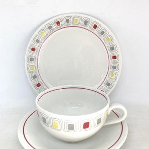 Puede incluir: Juego de té de cerámica blanca compuesto por una taza, un platillo y un plato. El juego presenta un patrón cuadrado rojo, gris y amarillo alrededor de los bordes. La taza tiene un asa y el plato es redondo.