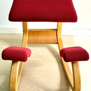 May include: A red kneeling chair with a wooden frame. The chair features a rectangular seat and two knee rests, all covered in red fabric. The wooden frame is light-coloured and curved.