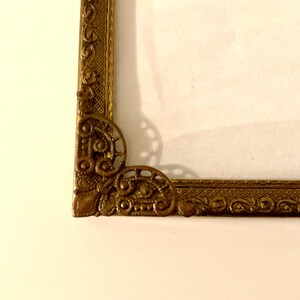 May include: Close-up of a decorative, ornate gold picture frame. The frame has intricate detailing in the corners and along the edges. The frame is set against a cream-colored background.