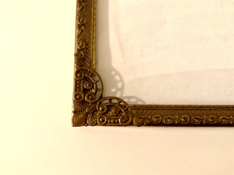 May include: Close-up of a decorative, ornate gold picture frame. The frame has intricate detailing in the corners and along the edges. The frame is set against a cream-colored background.