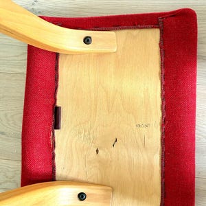 May include: A square, red upholstered chair seat with a wooden frame. The seat is attached to the frame with wooden supports and black fixings. The word "FRONT" is visible on the wooden seat.