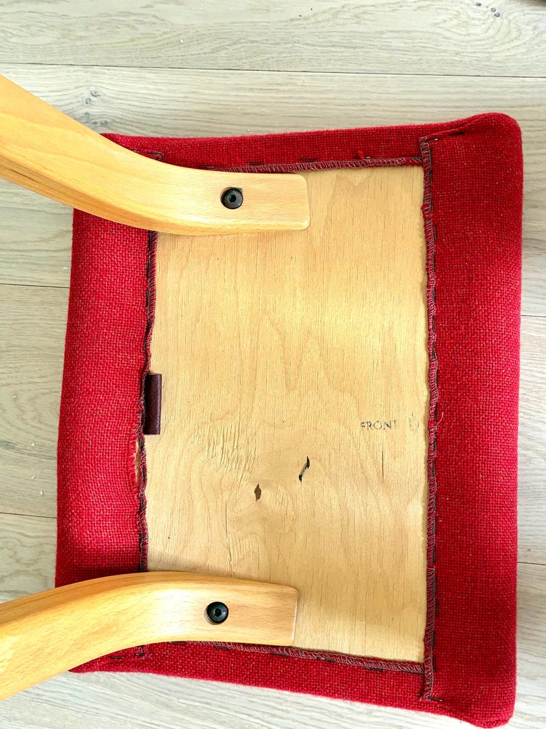 May include: A square, red upholstered chair seat with a wooden frame. The seat is attached to the frame with wooden supports and black fixings. The word "FRONT" is visible on the wooden seat.