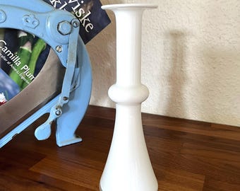 May include: A white, hourglass-shaped vase with a flared base and a wide, flat top. The vase has a bulbous midsection and stands approximately 8 inches tall. It is set against a wooden surface with a light blue object and a book in the background.