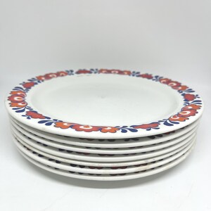 Puede incluir: Una pila de seis platos de cerámica blancos. Cada plato tiene un borde decorativo con un diseño floral en rojo, naranja y azul. Los platos están apilados uno encima del otro.