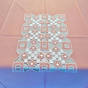 Puede incluir: Un mantel rosa con un centro bordado. El centro presenta un diseño geométrico en blanco y rosa, añadiendo un toque decorativo. El mantel está hecho de tela suave, ideal para la mesa.