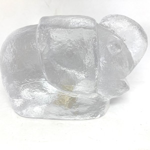 Puede incluir: Una figura de elefante de cristal transparente y texturizado. El elefante está en posición sentada con la trompa hacia abajo. La figura tiene detalles visibles de las orejas, los ojos y el cuerpo del elefante.