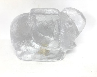 May include: A clear, textured glass elephant figurine. The elephant is in a seated position with its trunk down. The figurine has visible details of the elephant's ears, eyes, and body.