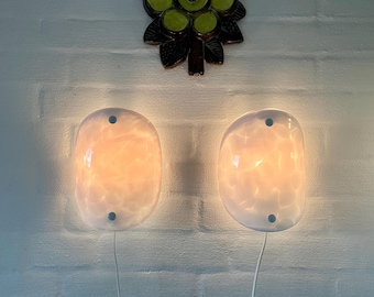 May include: Two oval wall sconces with white marbled glass shades and two small circular metal fasteners on each. The lamps emit a warm, soft glow and are mounted on a white brick wall. A dark brown ceramic flower decoration is positioned above.