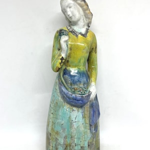 Puede incluir: Figura de cerámica de una mujer con un vestido largo, con la parte superior amarilla y la falda azul. La figura tiene un acabado agrietado y envejecido, con una cesta en las manos. La cabeza de la mujer está inclinada hacia abajo.