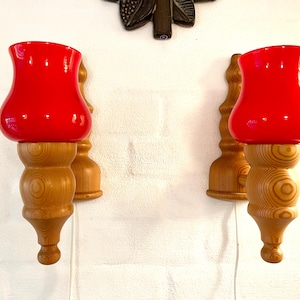 May include: A pair of wall sconces featuring red glass shades and wooden bases. The shades are a bright red, and the bases are crafted from light-coloured wood. The sconces are mounted on a white wall, with a decorative element above.
