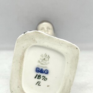 May include: The underside of a porcelain figurine base featuring a stamped Bing & Grondahl Copenhagen Denmark logo, the blue B&G mark, the number 1870, and the initials FL. The base is white with a recessed centre and a smooth, glazed finish.