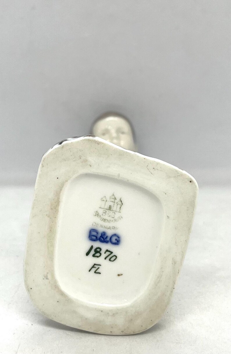 May include: The underside of a porcelain figurine base featuring a stamped Bing & Grondahl Copenhagen Denmark logo, the blue B&G mark, the number 1870, and the initials FL. The base is white with a recessed centre and a smooth, glazed finish.