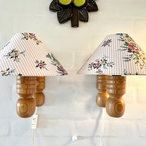 May include: A pair of wall-mounted lamps featuring light-colored wooden bases with a turned, cylindrical design. Each lamp has a conical fabric shade decorated with a pattern of pink vertical stripes and a colorful floral print of roses.