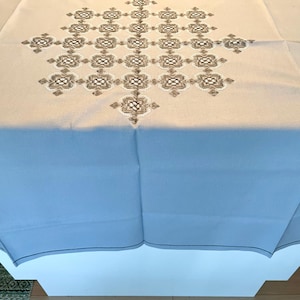 Puede incluir: Un mantel rectangular de color beige claro con un patrón geométrico bordado en el centro en tonos marrón y blanco. El diseño intrincado consiste en motivos cuadrados repetitivos dispuestos en forma de diamante sobre la tela.