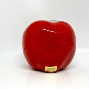 May include: A red ceramic apple shaped candle holder with a small opening on top for a tea light. The candle holder has a label on the bottom that reads "Rolf Berg, Tonarbeiten, Boden."