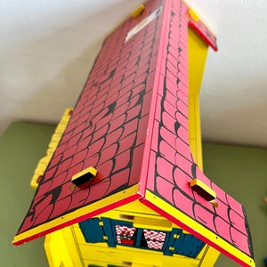 Op de afbeelding: Een houten speelgoedhuis met een rood dak en gele muren. Het dak heeft een zwart patroon en is geel afgewerkt. Het huis heeft blauwe luiken en kleine ramen. Het woord "MICKI" is zichtbaar aan de voorkant.