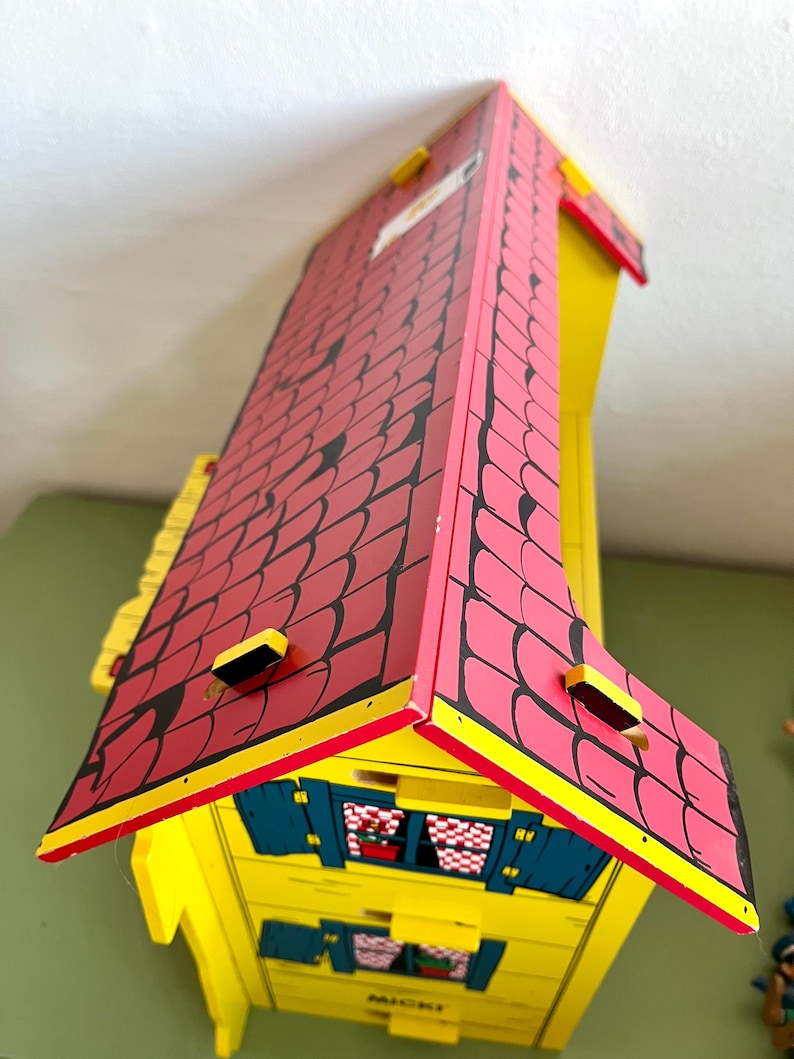 Op de afbeelding: Een houten speelgoedhuis met een rood dak en gele muren. Het dak heeft een zwart patroon en is geel afgewerkt. Het huis heeft blauwe luiken en kleine ramen. Het woord "MICKI" is zichtbaar aan de voorkant.