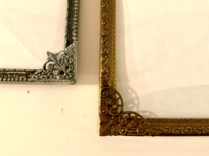 May include: Close-up of two ornate picture frame corners. One is silver with a textured finish, the other is gold with intricate scrollwork. Both frames have a vintage aesthetic and are set against a white background.